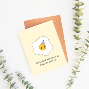 May include: A birthday card featuring a cartoon egg wearing a party hat. The card reads "HOPE YOUR BIRTHDAY IS EGGSTRA SPECIAL" on a pale yellow background, with a brown envelope behind it.