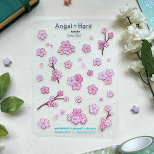 Può includere: Foglio di adesivi opachi trasparenti con illustrazioni di fiori di ciliegio rosa. Il foglio include fiori, rami e petali di varie dimensioni. Il testo "Angel + Hare" e "Sakura Sticker Sheet" è stampato in alto. Gli adesivi sono descritti come impermeabili.