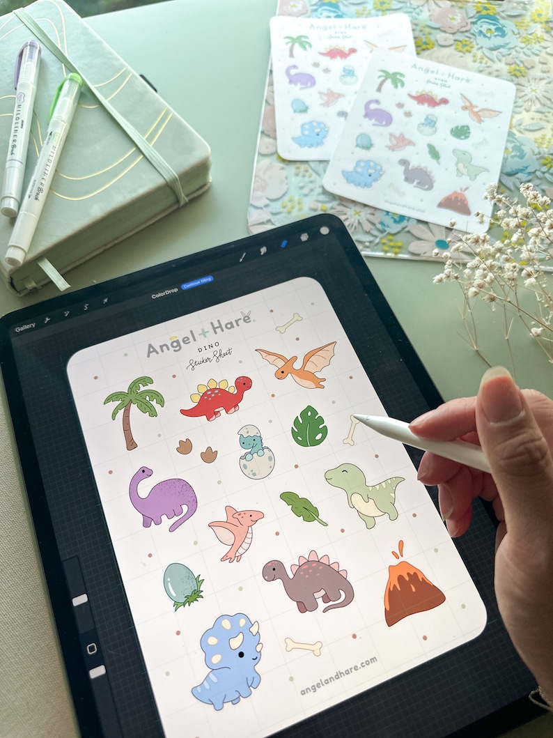 May include: A digital illustration of a sticker sheet featuring a variety of cute dinosaur designs. The sheet includes a variety of dinosaurs, including a triceratops, a stegosaurus, a pterodactyl, and a T-Rex. The sheet also includes a palm tree, a volcano, and a dinosaur egg. The sheet is titled "Angel + Hare Dino Sticker Sheet" and includes the website address "angelandhare.com".