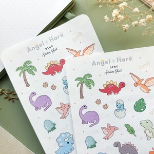 May include: Two sheets of colourful dinosaur stickers with a white background. The sticker sheets are labelled "Angel + Hare Dino Sticker Sheet". The stickers feature dinosaurs, palm trees, footprints, and bones.