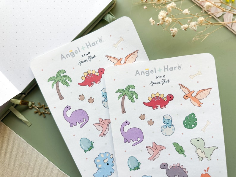 May include: Two sheets of colourful dinosaur stickers with a white background. The sticker sheets are labelled "Angel + Hare Dino Sticker Sheet". The stickers feature dinosaurs, palm trees, footprints, and bones.