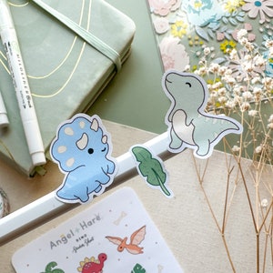 May include: Two cute dinosaur stickers, one blue triceratops and one green T-Rex, with a green leaf sticker. The stickers are on a white background with the text "Angel + Hare Dino Sticker Sheet" visible.