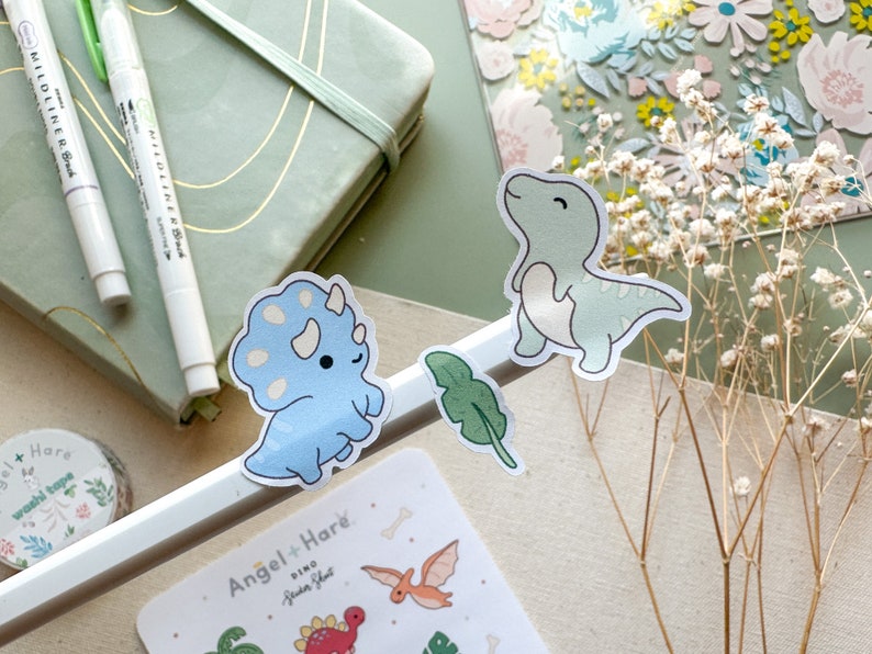 May include: Two cute dinosaur stickers, one blue triceratops and one green T-Rex, with a green leaf sticker. The stickers are on a white background with the text "Angel + Hare Dino Sticker Sheet" visible.