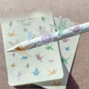 May include: A sheet of clear stickers with colorful origami crane designs. The stickers are arranged in a variety of sizes and positions on the sheet. The sheet is on a light pink surface.