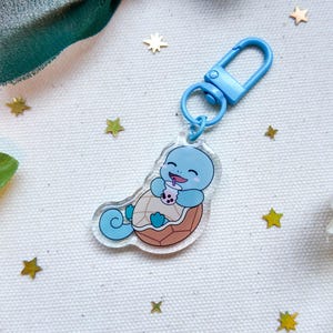 May include: A blue acrylic keychain featuring a cartoon turtle character holding a drink. The keychain has a blue clasp and ring. The turtle is light blue with a brown shell and is smiling. The background has gold star-shaped confetti.
