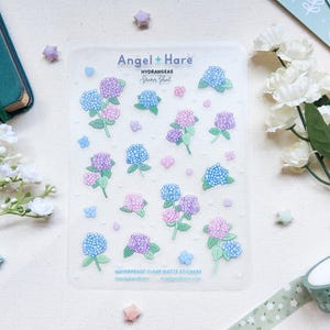 May include: A clear matte sticker sheet with illustrations of blue, purple, and pink hydrangeas. The sheet is labelled "Angel + Hare HYDRANGEAS Sticker Sheet". Small flower and star-shaped stickers are scattered around the sheet, along with a roll of floral washi tape.