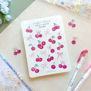 May include: A sticker sheet with illustrations of pink cherries and bows. The text "Angel + Hare Coquette Cherries Sticker Sheet" is visible. Two pink highlighters are also in the image. The stickers are on a white background.