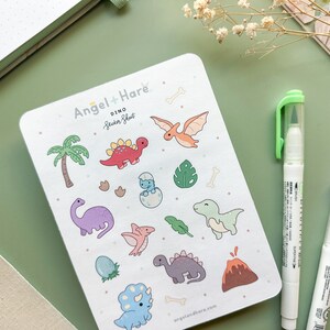 May include: A sticker sheet featuring a range of colourful dinosaur illustrations. The sheet is white with a pale green background and includes a palm tree, a volcano, and various dinosaur shapes. The sheet is labelled "Dino Sticker Sheet" and is from the brand "Angel + Hare".