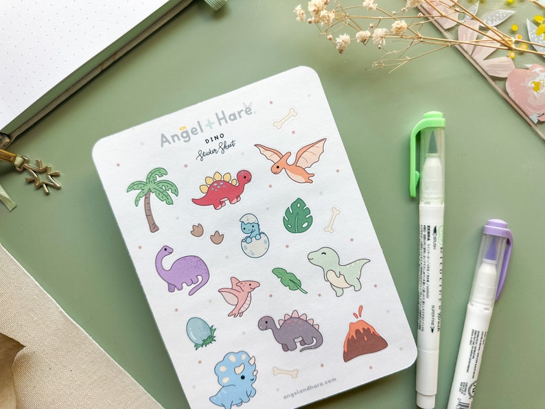 May include: A sticker sheet featuring a range of colourful dinosaur illustrations. The sheet is white with a pale green background and includes a palm tree, a volcano, and various dinosaur shapes. The sheet is labelled "Dino Sticker Sheet" and is from the brand "Angel + Hare".