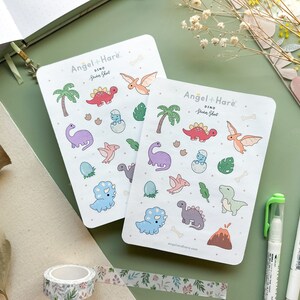 May include: Two sheets of colourful dinosaur sticker sheets with a white background. The sticker sheets are labelled "Dino Sticker Sheet" and feature a variety of dinosaurs, including a triceratops, stegosaurus, pterodactyl, and a T-Rex. The sticker sheets are from the brand "Angel + Hare."