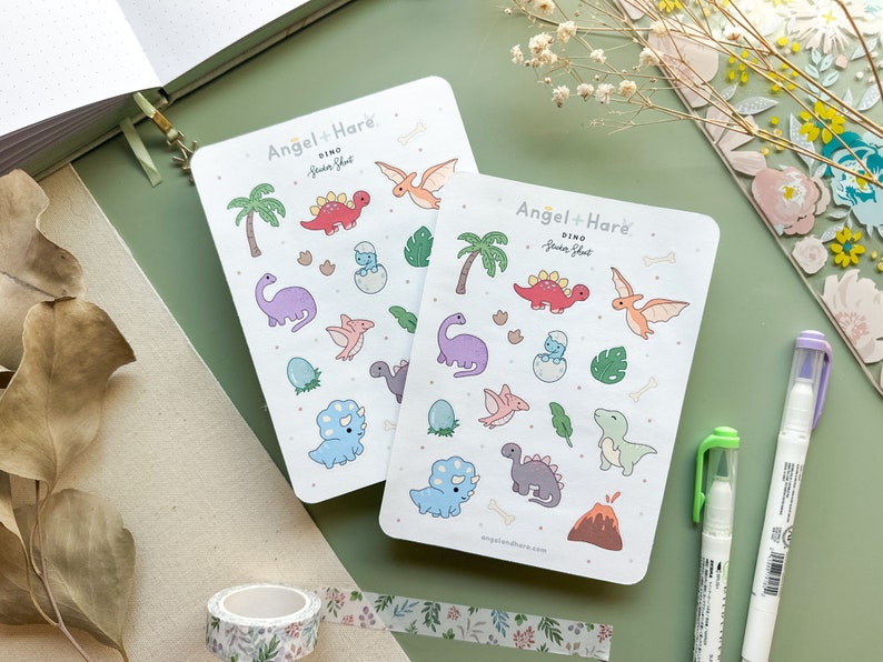 May include: Two sheets of colourful dinosaur sticker sheets with a white background. The sticker sheets are labelled "Dino Sticker Sheet" and feature a variety of dinosaurs, including a triceratops, stegosaurus, pterodactyl, and a T-Rex. The sticker sheets are from the brand "Angel + Hare."