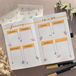 May include: A weekly planner with a January calendar and dates 18-24. The planner has a yellow and orange color scheme and features origami cranes hanging from the top of the page.