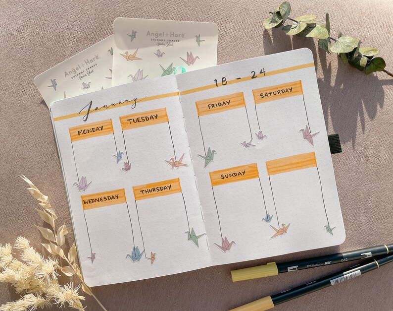 May include: A weekly planner with a January calendar and dates 18-24. The planner has a yellow and orange color scheme and features origami cranes hanging from the top of the page.