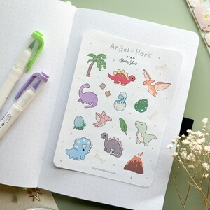 May include: A sheet of colourful dinosaur stickers featuring a variety of dinosaurs, including a T-Rex, a stegosaurus, and a pterodactyl. The stickers are on a white background with the text "Angel + Hare' Dino Sticker Sheet" at the top.