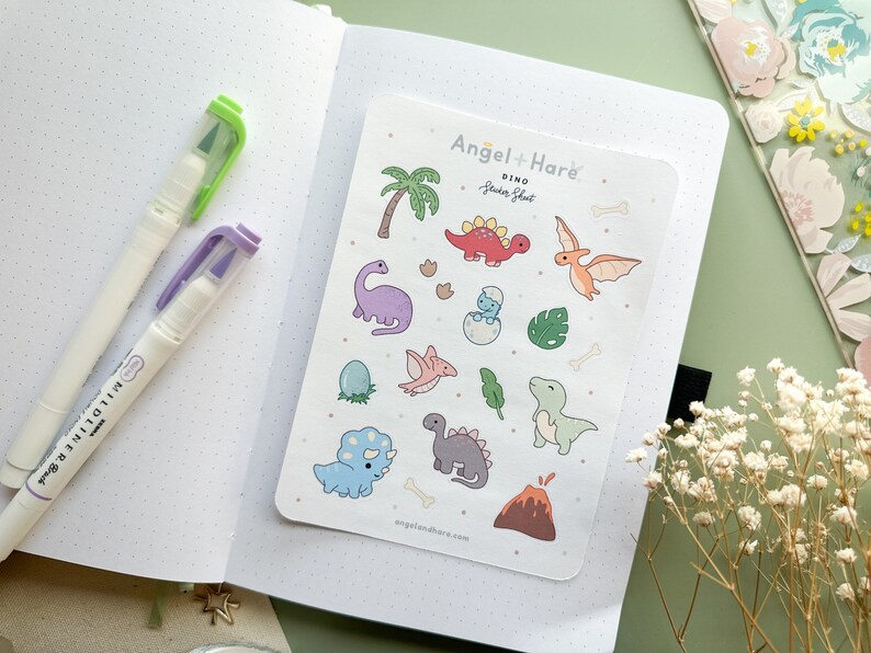May include: A sheet of colourful dinosaur stickers featuring a variety of dinosaurs, including a T-Rex, a stegosaurus, and a pterodactyl. The stickers are on a white background with the text "Angel + Hare' Dino Sticker Sheet" at the top.