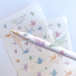 May include: A sheet of clear stickers with colorful origami crane designs. The stickers are on a white background and are being held by a pen.