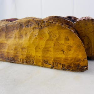 May include: A wooden sculpture of a Native American man wearing a headdress. The sculpture is painted in brown, red and yellow tones. The sculpture is signed "Vanguard Studios &copy; 1970".