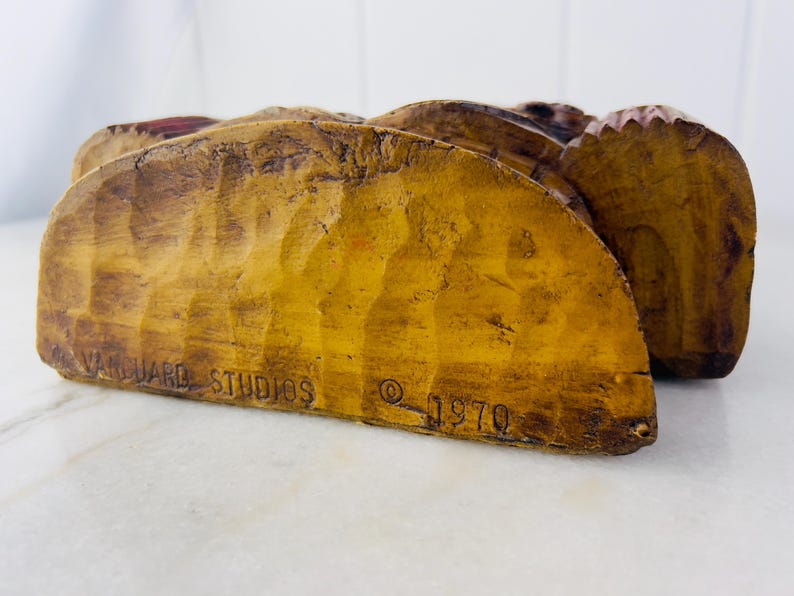May include: A wooden sculpture of a Native American man wearing a headdress. The sculpture is painted in brown, red and yellow tones. The sculpture is signed "Vanguard Studios &copy; 1970".