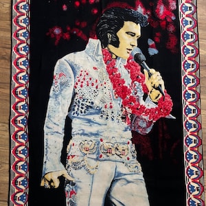 May include: A black tapestry featuring a portrait of Elvis Presley in a white jumpsuit with red and blue accents. The tapestry has a decorative border with red, white, and blue stars and stripes.