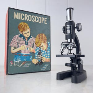 Puede incluir: Un microscopio negro con una lente plateada y una base negra. El microscopio tiene un aumento de 150x, 300x y 600x. El microscopio está en una caja con una imagen de dos niños usando un microscopio.
