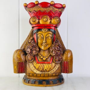 May include: A wooden sculpture of a woman wearing a red and gold crown and a long, flowing veil. The sculpture is painted in shades of brown, red, and gold.