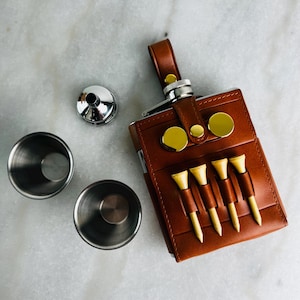 May include: A brown leather-wrapped metal flask with a top strap and gold-tone hardware, featuring four light-coloured wooden golf tees held in front slots. Included are two stainless steel shot cups and a small silver-tone metal funnel.