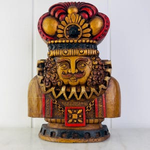 May include: A wooden figurine of a king with a red and gold crown, a gold beard, and a gold and red patterned chest. The figurine is painted in gold, red, and black.