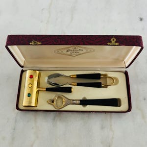 Könnte beinhalten: Ein Vintage-Bar-Werkzeugset in einer bordeauxroten Box. Das Set enthält einen goldfarbenen Eisbrecher mit roten und grünen Akzenten, einen Löffel, einen Flaschenöffner und ein kleines Werkzeug, alle mit schwarzen Griffen.