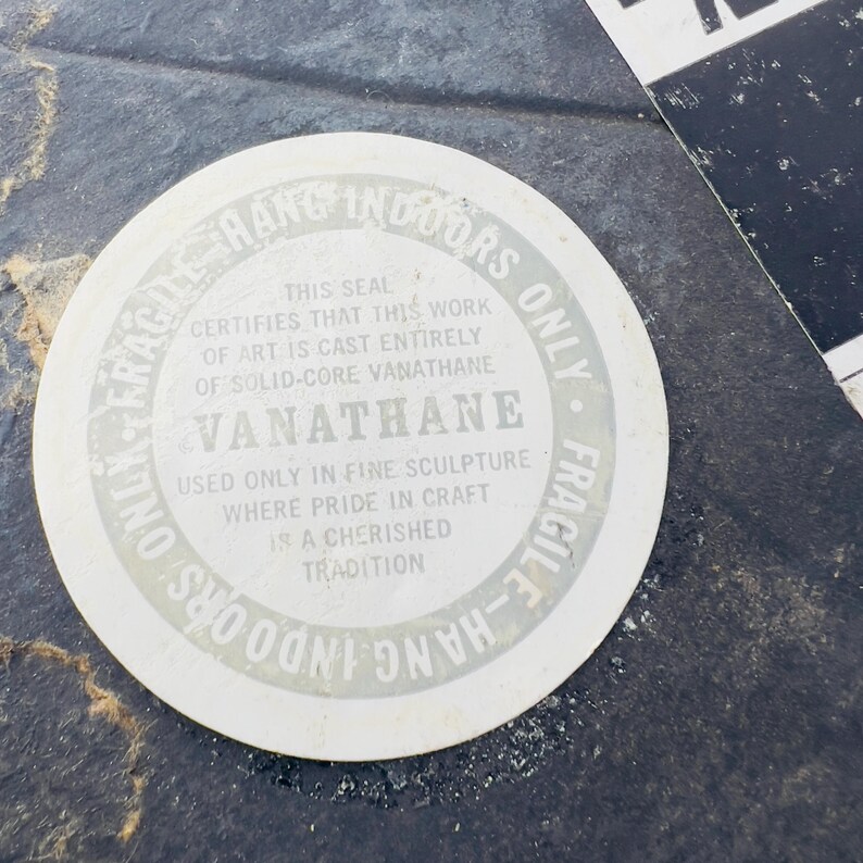 May include: A white round sticker with black text that reads "Fragile - Hang Indoors Only - Fragile - Hang Indoors Only". The sticker also reads "This seal certifies that this work of art is cast entirely of solid-core Vanathane Vanathane Used only in fine sculpture where pride in craft is a cherished tradition".