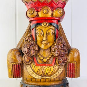 May include: A wooden bust of a woman wearing a red and gold crown and a long, flowing robe. The bust is painted in shades of gold, red, and black.