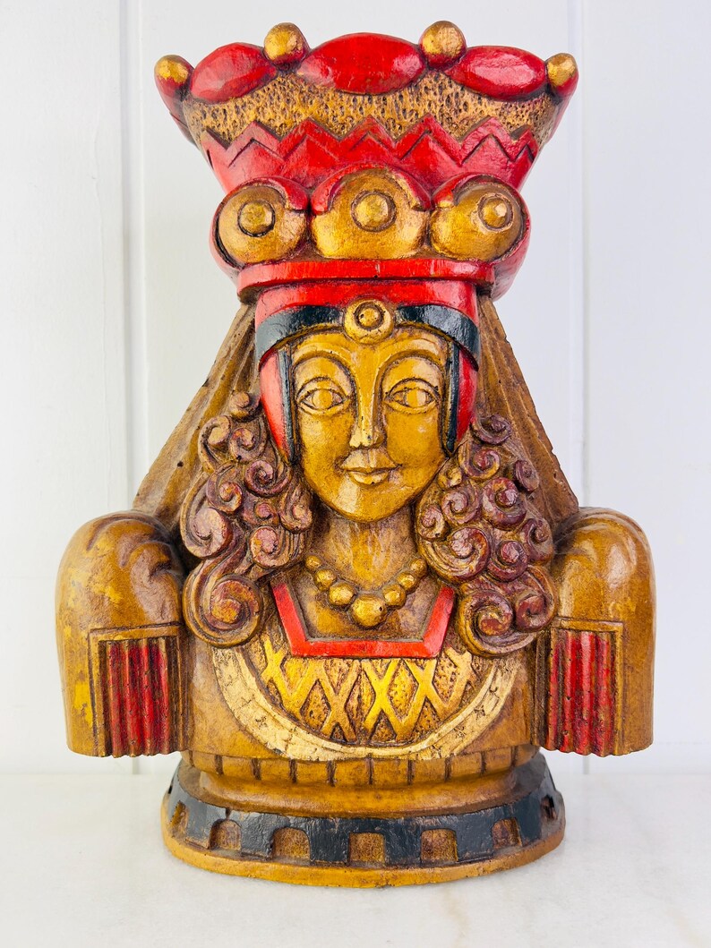 May include: A wooden bust of a woman wearing a red and gold crown and a long, flowing robe. The bust is painted in shades of gold, red, and black.