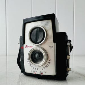 以下が含まれることがあります： Dakonレンズを備えた、黒とシルバーのヴィンテージBrownie Starflexカメラ。カメラには黒いストラップがあり、赤い文字で「Brownie」と書かれています。カメラには2つのレンズがあり、半円に「STARFLEX CAMERA」という文字があります。