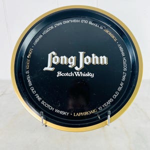 Könnte beinhalten: Ein runder, schwarzer Metalltablett mit goldenem Rand. Auf dem Tablett steht in weißer und goldener Schrift "Long John Scotch Whisky". Der Rand des Tabletts trägt die Aufschrift "Tormore 10 Years Old Highland Malt Scotch Whisky" und "Long John 12 Years Old Fine Scotch Whisky".