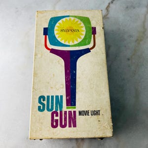 May include: A rectangular off-white cardboard box for a Sylvania Sun Gun movie light. The front features a graphic of a stylized light with a yellow sun icon, blue and green accents, and a purple handle. The text reads Sun Gun Movie Light.