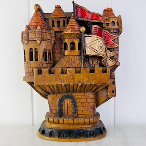 May include: A wooden castle figurine with a red and white flag. The castle is painted in shades of brown and orange, with a detailed brickwork design. The castle has a small door and windows.