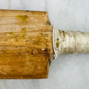 May include: Close-up of a vintage cricket bat. The wooden blade shows signs of wear with yellow-green stains. The handle is wrapped in white twine. The bat's design suggests it's an antique sports equipment.