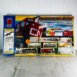 Könnte beinhalten: Ein Life-Like Trains Thundering Rails HO-Modelleisenbahn-Set in einer Schachtel mit einer rot-schwarzen Lokomotive Nummer 5506. Die Verpackung zeigt über 140 Teile, eine 114 cm mal 114 cm große Gleisanlage und verschiedene Güterwagen, darunter Texaco.