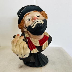 May include: A detailed bust of a clown with a black hat, black beard, and red and white scarf. The clown has a sad expression and is holding a cigar. The sculpture is painted with realistic details and is set on a marble surface.