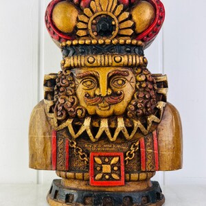 May include: A wooden figurine of a king with a red and gold crown, a gold chain, and a red and gold patterned chest. The figurine is painted in shades of gold, red, and black.