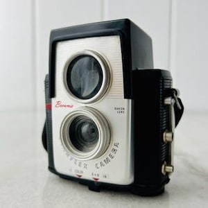 May include: A vintage Brownie Reflex camera with a black and silver body. The camera features two lenses, a red accent, and the text "Brownie" and "Reflex Camera". Includes a black strap and silver hardware.