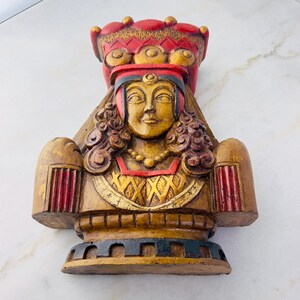 May include: A wooden sculpture of a woman wearing a crown and a red and gold robe. The sculpture is painted in shades of gold, red, and black.