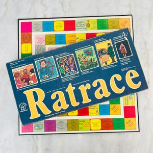 May include: A vintage Ratrace board game with a blue box and yellow lettering, resting on top of the game board. The board game has colorful squares and illustrations. The box reads "Ratrace" in large yellow letters.