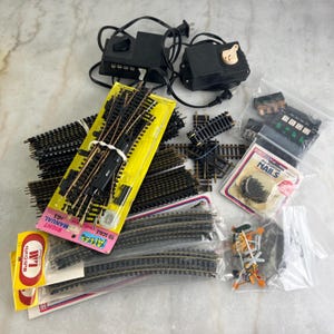 May include: A collection of model train accessories, including track pieces, a power supply, and small detail parts. The track is black with silver rails, and some pieces are still in their original packaging. The image includes the text "ATLAS HO SCALE".