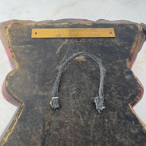 May include: The back of a red and black wall hanging with a gold plaque that reads "Wizard Studios, Inc. 1990". The hanging has two black cords attached to the back for hanging.