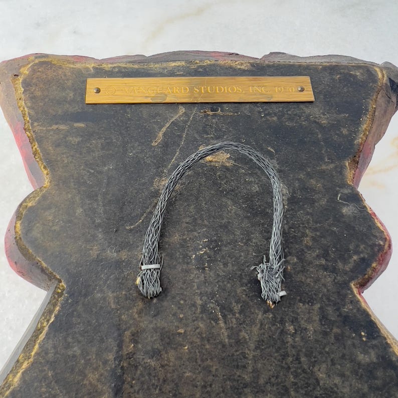 May include: The back of a red and black wall hanging with a gold plaque that reads "Wizard Studios, Inc. 1990". The hanging has two black cords attached to the back for hanging.