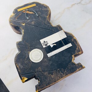 May include: A wooden wall sculpture of a stylised human figure with a dark brown and gold finish. The sculpture has a label on the back that reads "Anchored Studios" and "Yanatharee".