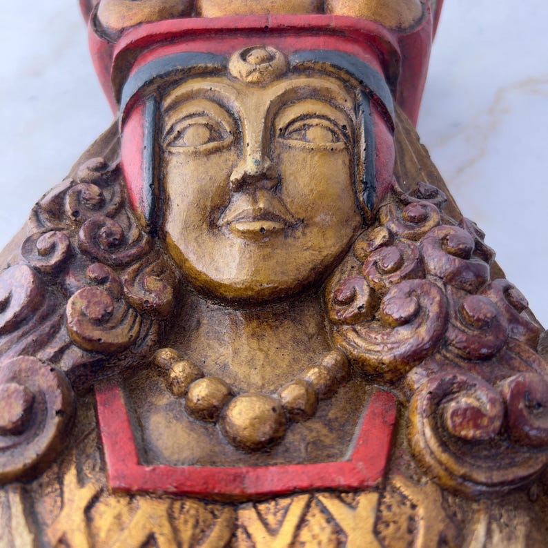 May include: A carved wooden sculpture of a woman's head with a red and gold crown. The woman has a detailed face with a gold necklace and curly hair.