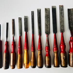 Könnte beinhalten: Eine Sammlung von zehn Holzmeißeln, nach Größe sortiert, mit Metallklingen und Griffen, die rot lackierte Oberteile und Unterteile aus Naturholz aufweisen. Die Werkzeuge variieren in der Breite, einige haben flache Klingen und eine ist als Hohleisen geformt.