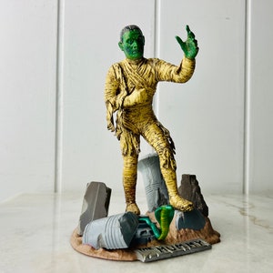 Peut inclure: Une figurine de momie au visage vert se tient sur une base avec des blocs de pierre en ruine et un serpent. La base est étiquetée "The Mummy".
