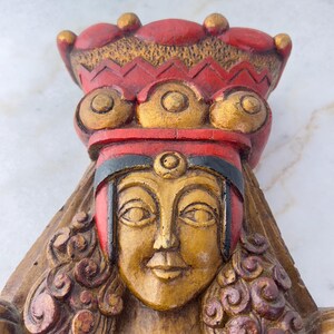 May include: A wooden sculpture of a woman's face with a red and gold crown. The woman's face is painted gold and has a serene expression. The crown is decorated with gold circles and has a red base.
